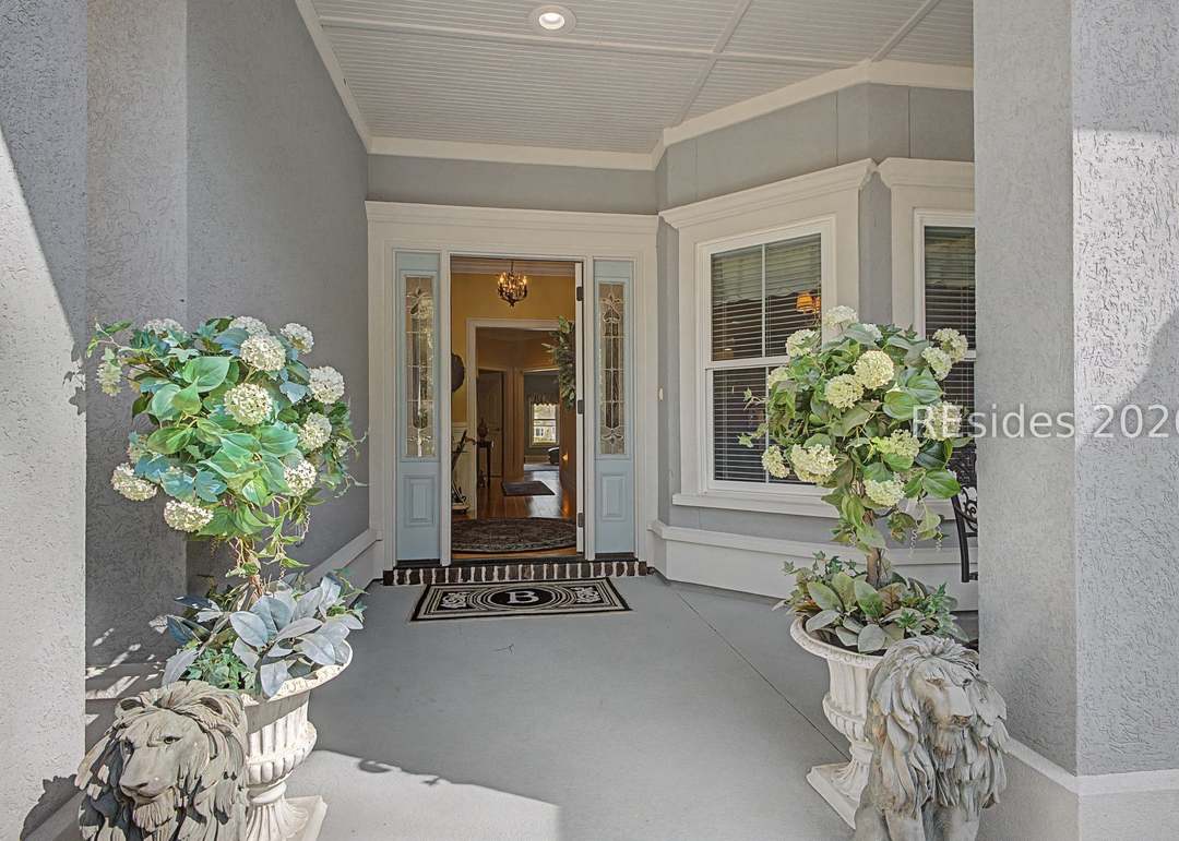 Covered front porch with twin columns, two large hydrangea topiaries flanking an open front door, a monogrammed welcome mat and lion-foot urns at either side