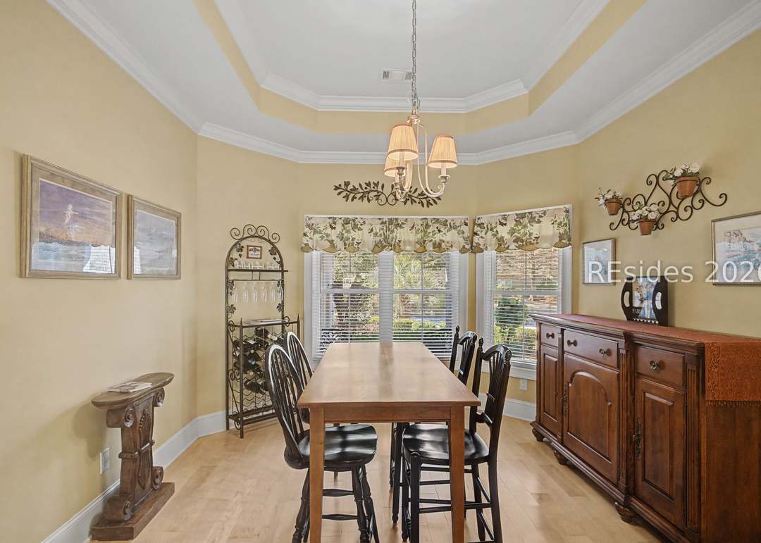Formal dining room with tray ceiling, crystal chandelier, long farmhouse table set for six and a tall bay window looking out to the side yard