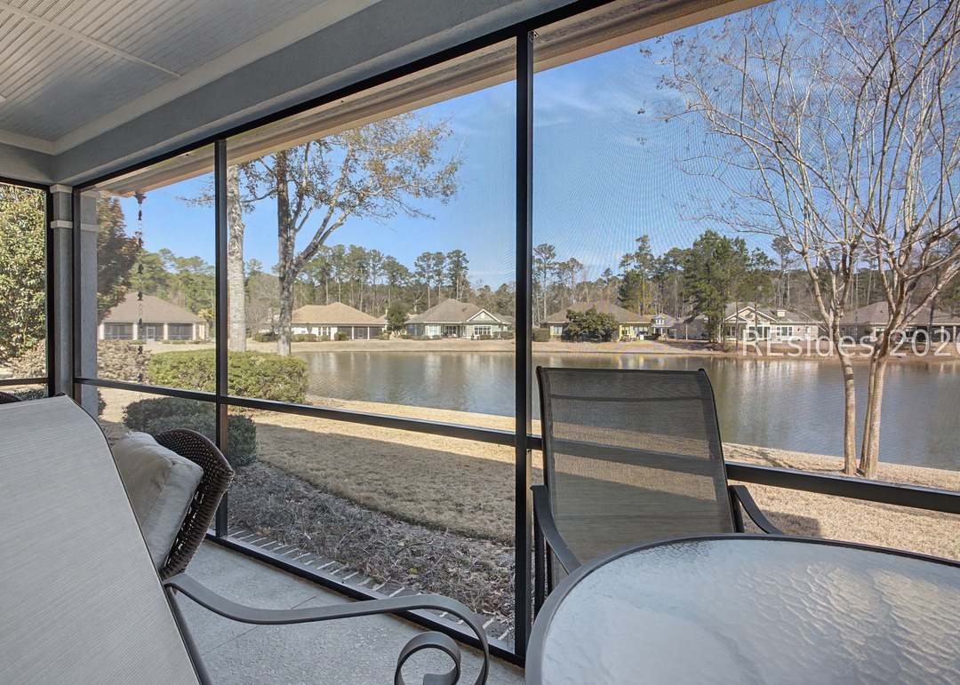 Chaise lounge angle of the screened lanai, looking out through the screen at the lagoon's curve, pine canopy and reflected sky